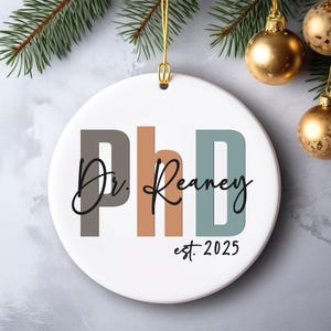 May include: White ceramic Christmas ornament with the text "PhD Dr. Reaney est. 2025" in gray, brown, and teal. Gold hanging string and ornaments are visible.
