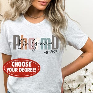 May include: Light gray t-shirt with the text "PharmD Dr. Guevara est. 2026" in a mix of colors. A red oval sticker at the bottom says "CHOOSE YOUR DEGREE!"