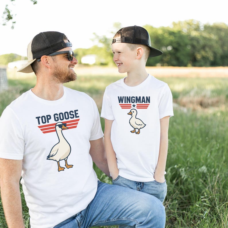 May include: Two white t-shirts with matching designs. One shirt says "TOP GOOSE" with a cartoon goose wearing sunglasses. The other says "WINGMAN" with a cartoon duck. Both shirts have red and blue stripes.