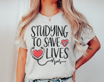 May include: A light gray short-sleeve t-shirt featuring the black text "STUDYING TO SAVE LIVES" with a red heart inside a black stethoscope graphic and a small red heart with a white EKG line. The design is centered on the front of the shirt.