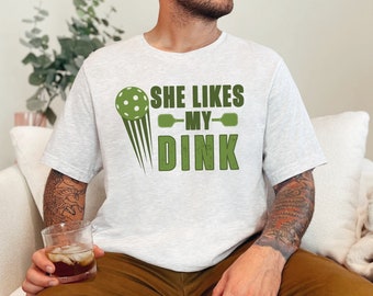 Op de afbeelding: Lichtgrijs t-shirt met de tekst "SHE LIKES MY DINK" in het groen. Het ontwerp bevat een pickleball en paddles. Het shirt is gemaakt van een zacht materiaal.