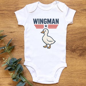 May include: White baby onesie with the word "WINGMAN" in navy blue block letters above a cartoon duck. The design includes red and white stripes and a white star. The onesie is on a wooden surface.
