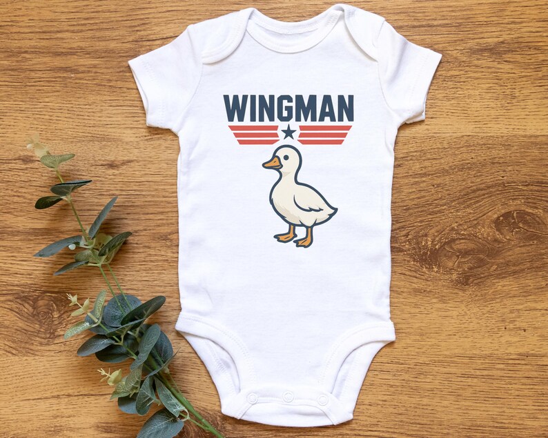 May include: White baby onesie with the word "WINGMAN" in navy blue block letters above a cartoon duck. The design includes red and white stripes and a white star. The onesie is on a wooden surface.