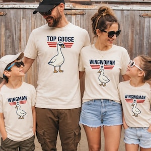May include: A family wearing matching beige t-shirts with a goose graphic and text. The shirts say "Top Goose," "Wingwoman," and "Wingman." The goose graphic has sunglasses. The family is standing in front of a wooden fence.