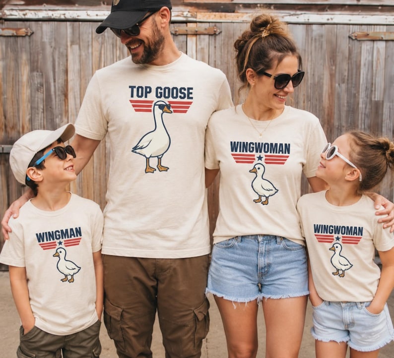 May include: A family wearing matching beige t-shirts with a goose graphic and text. The shirts say "Top Goose," "Wingwoman," and "Wingman." The goose graphic has sunglasses. The family is standing in front of a wooden fence.