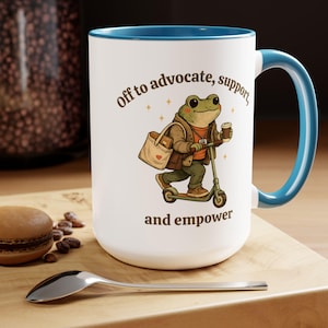May include: White ceramic mug with a blue interior and handle. The mug features a cartoon frog riding a scooter, carrying a tote bag and coffee cup. Text on the mug reads "Off to advocate, support, and empower."