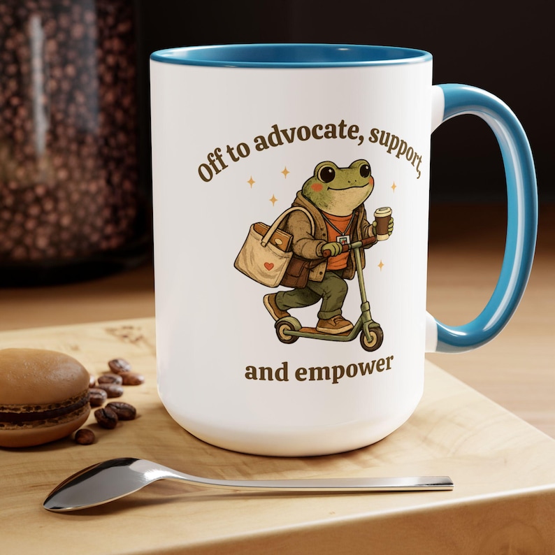 May include: White ceramic mug with a blue interior and handle. The mug features a cartoon frog riding a scooter, carrying a tote bag and coffee cup. Text on the mug reads "Off to advocate, support, and empower."