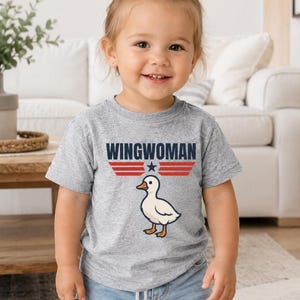 May include: A toddler wearing a gray t-shirt with the text "WINGWOMAN" above a cartoon duck illustration. The text is in navy blue with red stripes and a star. The child is also wearing denim shorts.