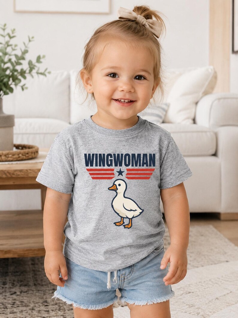 May include: A toddler wearing a gray t-shirt with the text "WINGWOMAN" above a cartoon duck illustration. The text is in navy blue with red stripes and a star. The child is also wearing denim shorts.
