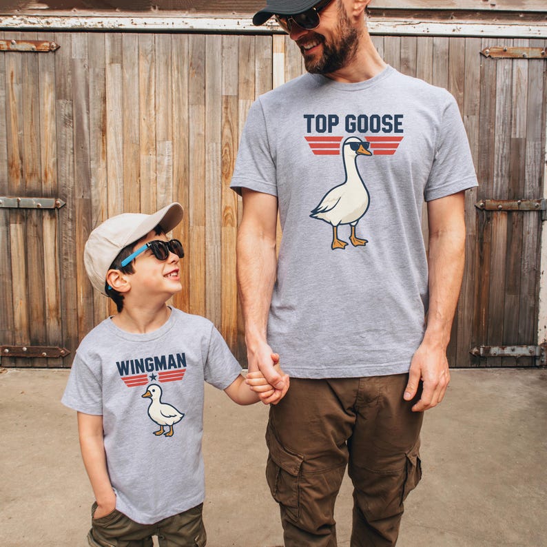 May include: Two gray t-shirts with matching designs. One shirt says "TOP GOOSE" with a goose wearing sunglasses. The other shirt says "WINGMAN" with a matching goose. The shirts are worn by a parent and child.