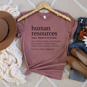 May include: Mauve t-shirt with the words "human resources" and its definition printed in black. The definition includes the unofficial lawyer, psychologist, event planner, teacher, peace maker, career planner, and detective. The shirt is displayed with a brown hat, jeans, and shoes.
