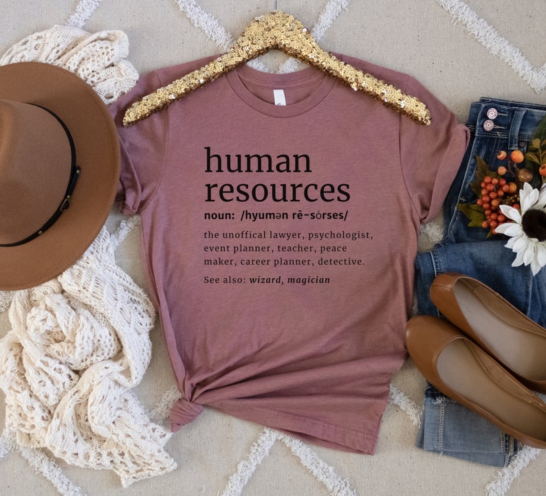 May include: Mauve t-shirt with the words "human resources" and its definition printed in black. The definition includes the unofficial lawyer, psychologist, event planner, teacher, peace maker, career planner, and detective. The shirt is displayed with a brown hat, jeans, and shoes.