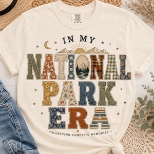 May include: Cream-colored t-shirt with the text "IN MY NATIONAL PARK ERA" in a patchwork design. The design includes a mountain scene, stars, and the phrase "COLLECTING CAMPSITE MEMORIES".