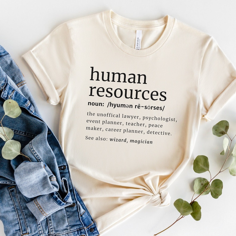 May include: Cream-colored t-shirt with the words "human resources" printed in black, along with a definition and related professions. The shirt is folded and placed on top of a denim jacket.
