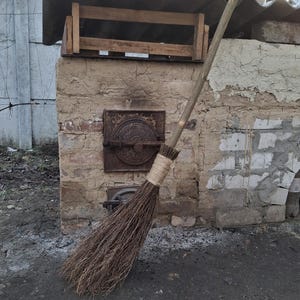 May include: A traditional brown broom with a long wooden handle resting against a brick wall. The broom is made from natural materials and has a rustic look.
