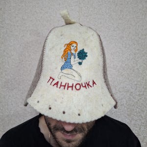 May include: A white felt hat with a brown brim and a red embroidered design of a woman holding a bouquet of flowers. The text "ПАННОЧКА" is embroidered in red on the front of the hat.