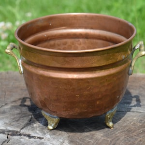 Könnte beinhalten: Ein Kupfertopf mit Messinggriffen und Füßen. Der Topf hat eine gehämmerte Oberfläche und steht auf einer Holzoberfläche.
