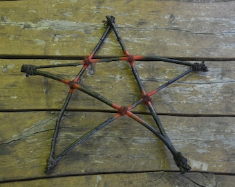 May include: A rustic, handmade pentagram crafted from dark, slender branches. The star's points are joined with red-orange twine, and the branches are tied together with dark twine. The star is set against a weathered, wooden background.