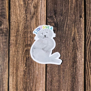 May include: A sticker featuring a grumpy cartoon seal with the text "I DON'T GIVE A" in colorful letters. The seal is gray with a white outline and is set against a wooden background.