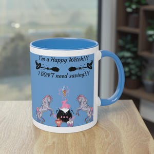 Op de afbeelding: Witte keramische mok met blauw interieur en handvat. De mok heeft de tekst "I'm a Happy Witch!!! I DON'T need saving!!!" met illustraties van eenhoorns, een ketel en hekserij-elementen. Op een houten oppervlak.