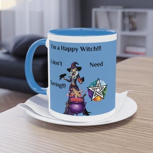 Op de afbeelding: Een keramische mok met een blauwe binnenkant en handvat, met een heksenillustratie en de tekst "I'm a Happy Witch!!! I don't Need Saving!!!". De heks wordt afgebeeld met een raaf, ketel en pentagram symbool.