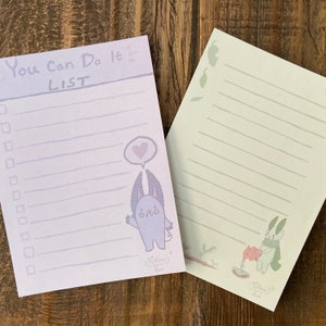 Könnte beinhalten: Zwei linierte Notizblöcke in Lila und Grün mit einem niedlichen Monster-Cartoon und einem Hasen auf jedem. Der Notizblock links hat den Text "You Can Do It List" und der Notizblock rechts hat den Text "Chibi Love & Beast".