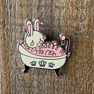 May include: A white bunny with pink ears is taking a bath in a white bathtub with pink bubbles. The bathtub has a blue faucet and blue flowers on the side.