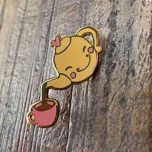 Puede incluir: Pin de esmalte dorado de una tetera de dibujos animados vertiendo té en una taza. La tetera tiene una cara sonriente y mejillas rosadas. La taza es rosa.