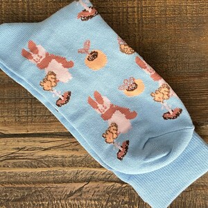 Könnte beinhalten: Ein Paar hellblaue Socken mit einem sich wiederholenden Muster aus braunen und rosa Hasen, Blumen und anderen dekorativen Elementen. Die Socken sind aus einem weichen, bequemen Material gefertigt.