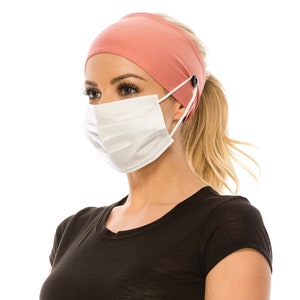 May include: A woman wearing a coral headband with buttons to hold a face mask in place. The headband is made of a soft, stretchy fabric. The woman is wearing a black t-shirt.