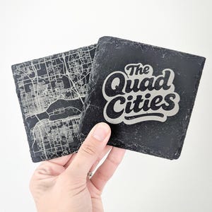 May include: Two square black slate coasters with rough, natural edges. One coaster features a detailed white engraved street map, while the other displays the white text The Quad Cities in a bold, stylized, rounded script font.