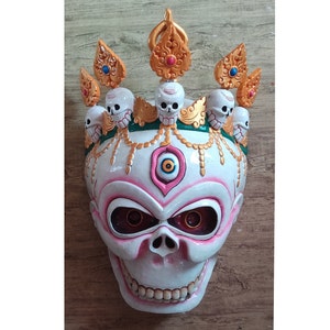 May include: A white skull mask with a pink third eye and gold accents. The mask is decorated with a crown of small skulls and gold ornaments.