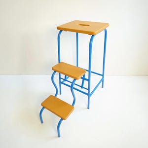 May include: A three-step stool featuring a bright blue tubular metal frame and three rectangular mustard yellow wooden steps. The top step includes a rectangular cutout handle, and the frame is constructed with curved supports for the lower steps.
