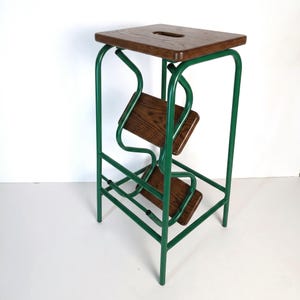 Op de afbeelding: Een groene metalen en houten opstap. De kruk heeft een vierkante, donkere houten bovenkant met een handgreepuitsparing. Twee houten treden worden ondersteund door een groen metalen frame. Het frame heeft een uniek, gebogen ontwerp.