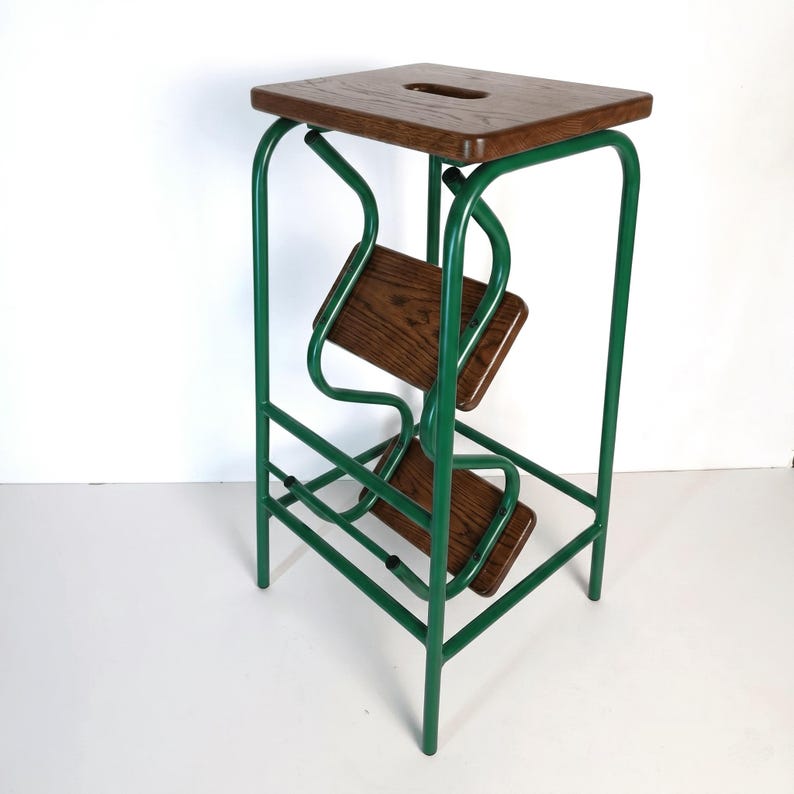 Op de afbeelding: Een groene metalen en houten opstap. De kruk heeft een vierkante, donkere houten bovenkant met een handgreepuitsparing. Twee houten treden worden ondersteund door een groen metalen frame. Het frame heeft een uniek, gebogen ontwerp.
