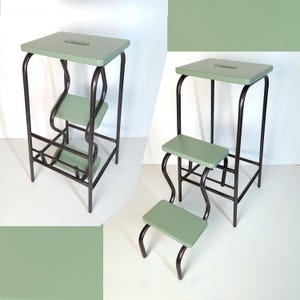 Könnte beinhalten: Ein salbeigrüner Tritthocker mit drei Stufen. Der Hocker hat eine rechteckige Oberseite mit einem Griffausschnitt, zwei untere Stufen und einen dunklen Metallrahmen. Der Rahmen hat gebogene Stützen für die Stufen und gerade Beine.