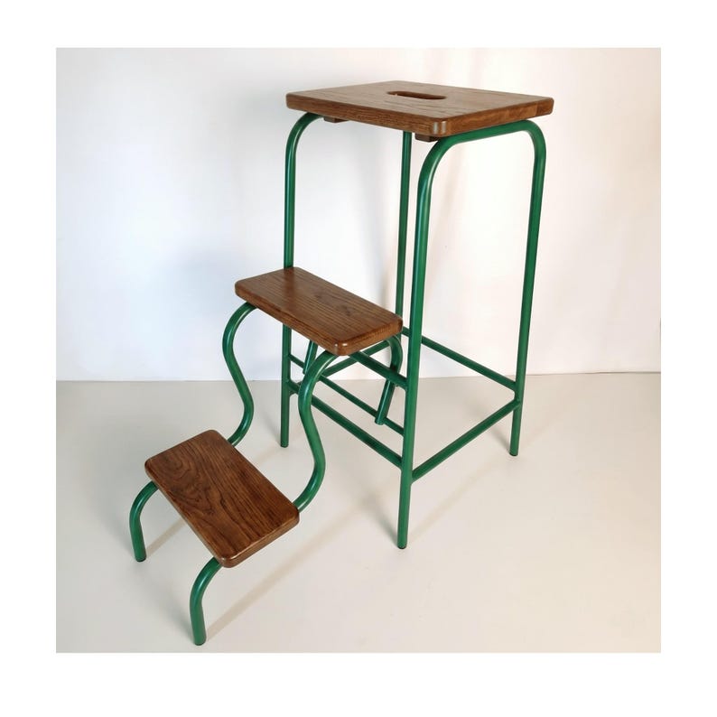 Op de afbeelding: Een vintage opstapje met drie houten treden en een groen metalen frame. De bovenste trede is rechthoekig met een uitsparing en de onderste treden hebben een gebogen ontwerp. De kruk is ontworpen om hogere plaatsen te bereiken.