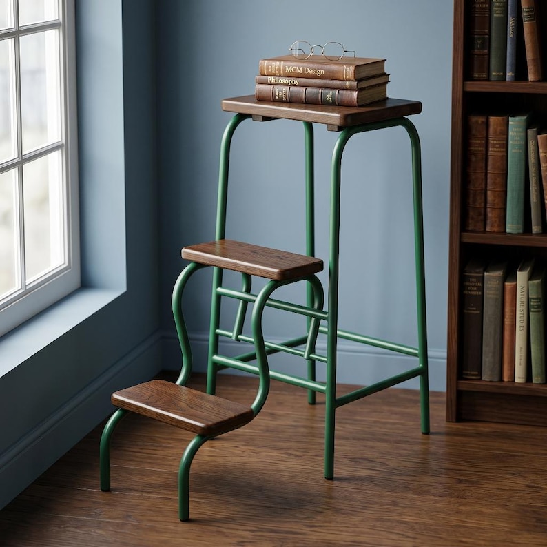Op de afbeelding: Een drietraps kruk met een groen metalen frame en houten treden. Op de bovenste trede liggen twee boeken en een bril. De treden hebben een donkere houten afwerking en het frame is groen geschilderd. Op de achtergrond een boekenkast en een raam.