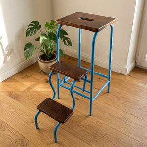 May include: A three-step stool featuring a bright blue tubular metal frame and three dark brown wooden steps. The top step includes a rectangular cutout handle, and the frame is constructed with curved supports connecting the steps to the base.