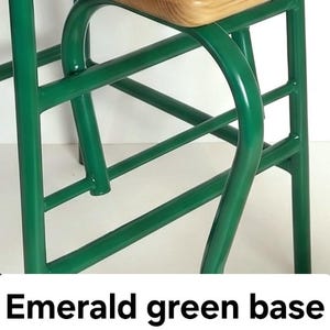 Op de afbeelding: Close-up van een stoelbasis met een smaragdgroen metalen frame. Het frame heeft gebogen en rechte lijnen, met een lichtgekleurde houten zitting aan de bovenkant. De tekst "Emerald green base" staat onderaan.