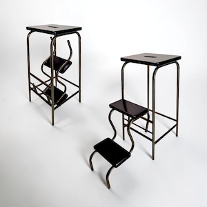 May include: Two black and gold metal step stools. Each stool has a square top with a cutout, a lower step, and a curved, decorative frame. The stools are on a white background.