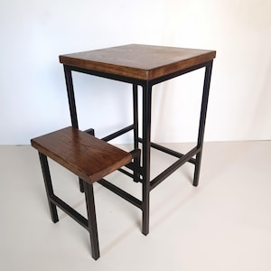 May include: A tall square table with a dark brown wood top and a black metal frame, featuring an attached lower step or footrest also made of dark brown wood with a matching black metal frame, creating a two-tiered structure for seating.