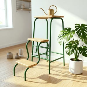 Puede incluir: Un taburete de metal y madera verde con tres escalones. Una pequeña regadera de latón se encuentra en el escalón superior. A la derecha del taburete hay una planta en maceta con grandes hojas verdes. La habitación está bien iluminada.