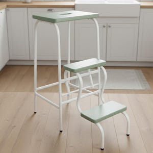 Puede incluir: Un taburete blanco y verde con una parte superior rectangular y dos escalones inferiores. El taburete tiene un diseño minimalista con un marco de metal blanco y escalones verdes. El escalón superior tiene un pequeño recorte.