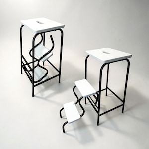 Puede incluir: Tres taburetes modernos con asientos blancos y marcos de metal negro. Los taburetes tienen un diseño minimalista, con una parte superior rectangular y un asa recortada. Un taburete tiene dos escalones, otro uno y el tercero un solo escalón.