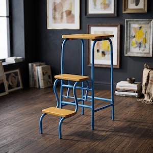 Op de afbeelding: Een trapje met drie treden voorzien van een felblauw buisvormig metalen frame en drie platte, mosterdgele houten treden. De bovenste trede heeft een kleine ronde uitsparing en de constructie wordt ondersteund door gebogen metalen poten.