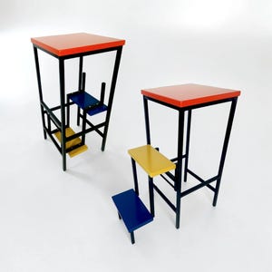 Puede incluir: Dos taburetes modernos y geométricos. Cada taburete tiene una parte superior cuadrada roja, un marco negro y un escalón azul y amarillo. Los taburetes están sobre un fondo blanco.