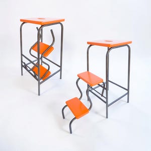 Op de afbeelding: Twee moderne krukjes met oranje zittingen en een donker metalen frame. De krukjes hebben een vierkante bovenkant met een gat en twee of drie oranje treden. Het metalen frame heeft een uniek, gebogen ontwerp.