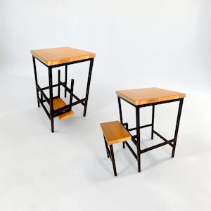 Puede incluir: Dos taburetes de madera y metal. Los taburetes tienen una tapa de madera de color claro y una estructura de metal oscuro. Un taburete tiene un reposapiés. Los taburetes están sobre un fondo blanco.