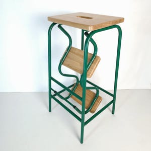 Op de afbeelding: Een groene metalen en houten opstap met een vierkante bovenkant en twee lagere treden. Het frame is heldergroen en de treden en bovenkant zijn van lichtgekleurd hout. De kruk heeft een uniek, modern ontwerp.